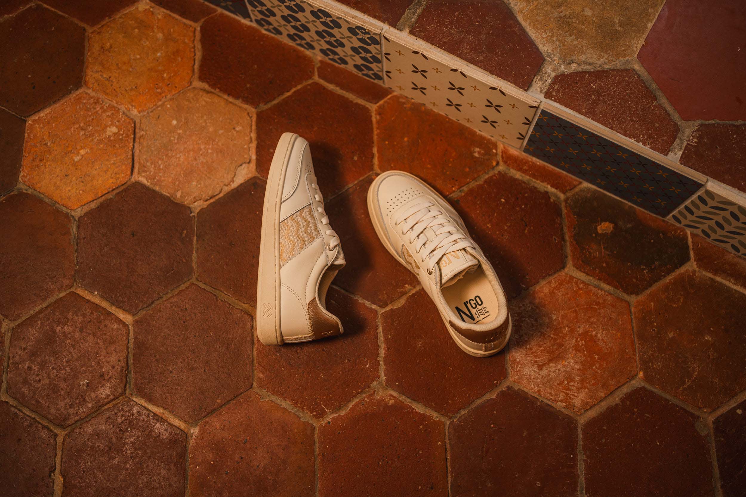 Zapatillas ecológicas hechas con materiales naturales, colgadas del techo por sus cordones, creando una instalación aérea bajo una viga de madera rústica. Estas zapatillas sostenibles, elaboradas con tela tejida artesanal y cuero vegano, están disponibles